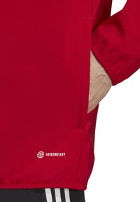 Rode sportjack met een gladde textuur, elastische manchetten, zijzak en een wit "AEROREADY" logo aan de onderkant.
