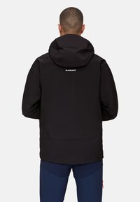 Giacca impermeabile nera con cappuccio, caratterizzata da una finitura testurizzata e un logo bianco sulla parte alta della schiena. Abbinata a pantaloni blu scuro.