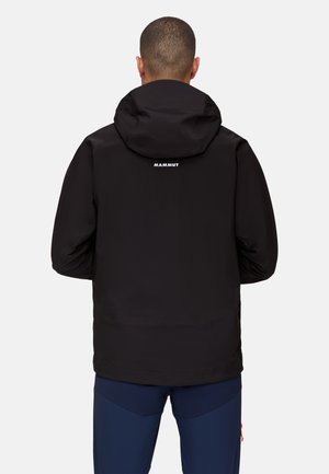 Schwarze wasserdichte Jacke mit Kapuze, ausgestattet mit einer strukturierten Oberfläche und einem weißen Logo auf dem oberen Rücken. Kombiniert mit dunkelblauen Hosen.