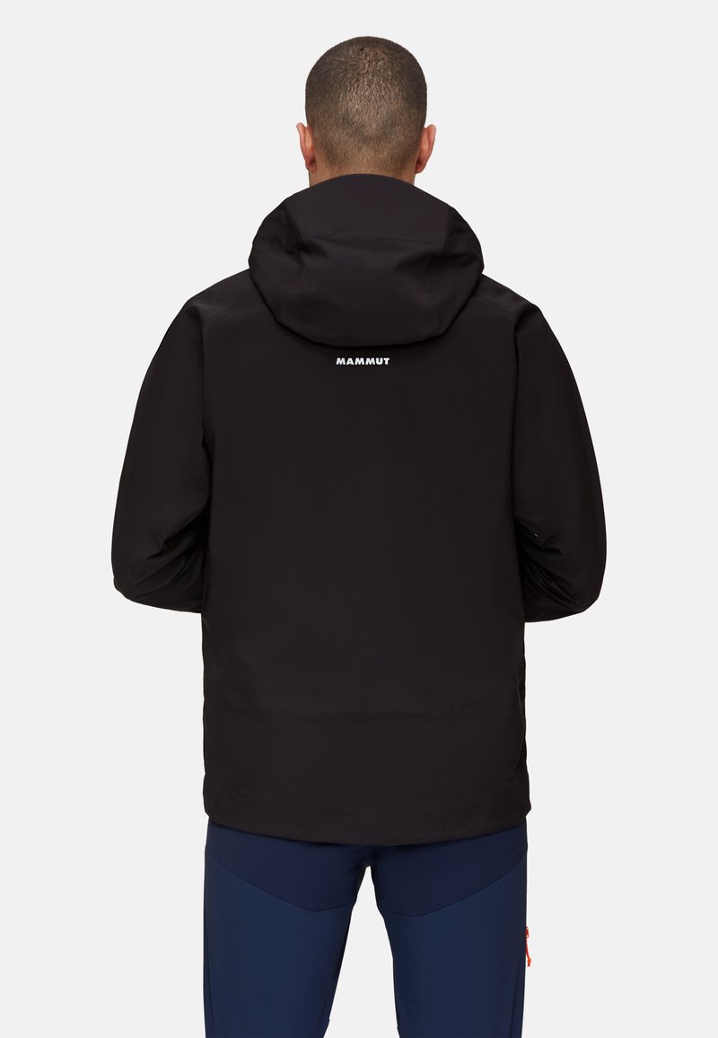 Giacca impermeabile nera con cappuccio, caratterizzata da una finitura testurizzata e un logo bianco sulla parte alta della schiena. Abbinata a pantaloni blu scuro.