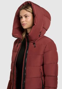 Chaqueta acolchada roja con capucha, cuello alto, cierre de cremallera negro y cordón. Textura suave con diseño de paneles cosidos.