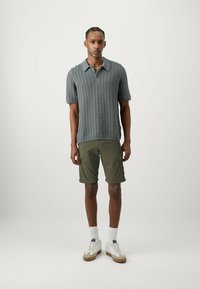 Polo grigio a maglia con un motivo a trame, maniche corte e colletto; pantaloncini cargo olivastri; calze bianche; sneaker bianche con suole marroni.