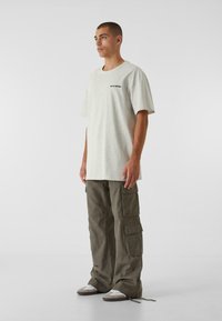 Lys grå bomulds T-shirt med korte ærmer, der har et lille sort trykt logo. Olivengrøn cargobukser med flere lommer og afslappet pasform.