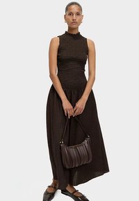 Hnědý texturovaný top bez rukávů s imitací roláku, doplněný dlouhou černou sukní a žebrovanou hnědou kabelkou. Modelka má na nohou černé baleríny.