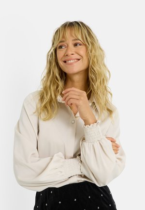 Camisa de manga larga beige claro con cierre de botones y puños elásticos con volantes; tela lisa; combinada con una falda negra texturizada.