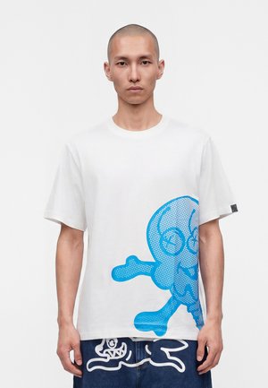 Mand med barberet hoved iført en hvid t-shirt med et stort blåt tegneseriekranie og korsben på nederste højre side.