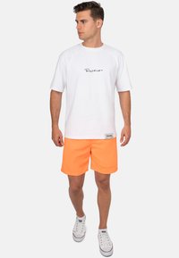 T-shirt di cotone bianca con logo nero, abbinata a pantaloni corti arancioni e sneakers bianche. Stile casual, design semplice e colori vivaci.