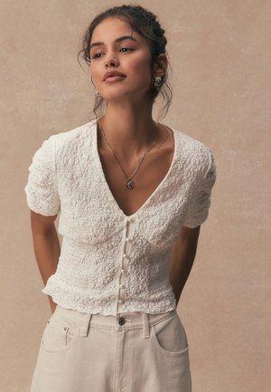 Žena v texturované bílé krátké haleně na knoflíky s perlovými knoflíky a béžových kalhotách s vysokým pasem pózuje na neutrálním pozadí.