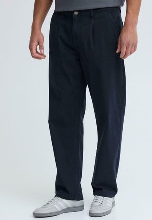 Homme portant un pantalon noir à coupe décontractée et des baskets blanches, debout devant un fond clair uni.