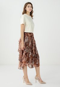 Crème korthangend mouwtop gecombineerd met een bruine rok met paisleypatroon en meerdere lagen. Neutraal gekleurde hoge hakken passen goed bij de outfit.