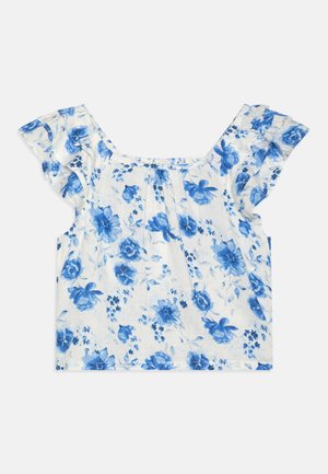 Hvid bluse med korte ærmer med blåt blomsterprint og flæser på skulderen, lagt fladt på en neutral baggrund.