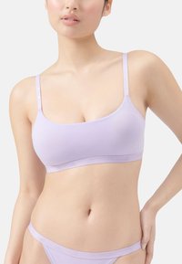 Frau in einem helllila nahtlosen BH und passender Unterwäsche, die mit einer Hand auf der Hüfte gegen einen schlichten Hintergrund steht.