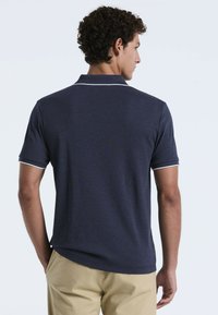 Polo shirt bleu marine en tissu texturé, avec un col blanc et des accents sur les manches, à manches courtes et une coupe décontractée. Design décontracté, vue arrière montrée.