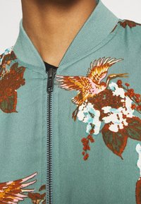 Chaqueta bomber de color azulado con bordado floral que presenta pájaros, rojos y rosas vibrantes, cierre con cremallera y detalle de cuello acanalado.