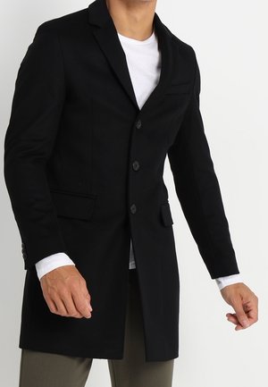 Homme portant un manteau noir boutonné par-dessus une chemise blanche et un pantalon vert olive, debout devant un fond blanc uni.