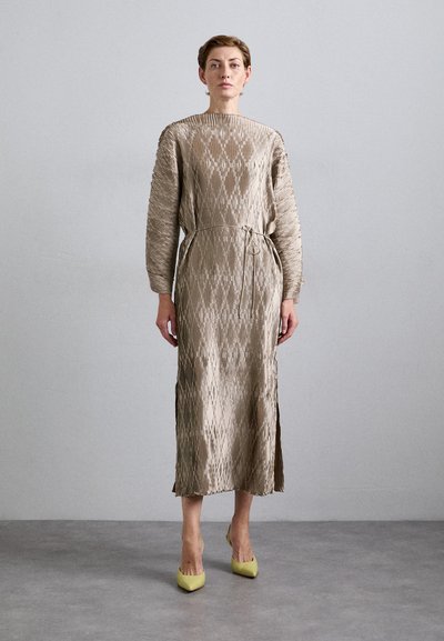 Femme debout contre un mur gris, portant une longue robe taupe texturée à manches longues et des escarpins jaunes à bout pointu.