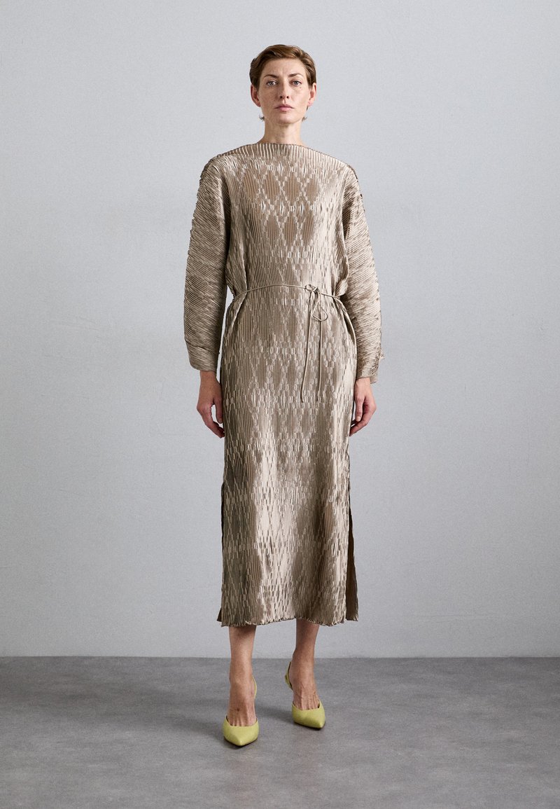 Woman standing against gray wall wearing a long, textured taupe dress with long sleeves and yellow pointed-toe heels.
