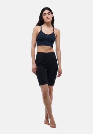 Jeune femme debout pieds nus portant un soutien-gorge de sport bleu marine foncé et un short cycliste noir taille haute sur un fond clair uni.