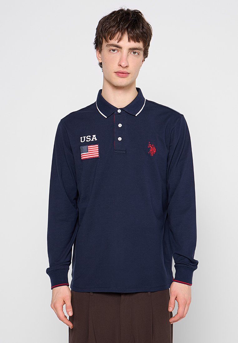 Polo de manga larga azul marino con cuello blanco, con acentos rojos, emblema de la bandera de EE. UU. y logo en el pecho. Material de algodón suave.