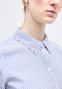 Κοντινό πλάνο ατόμου που φορά ασημένια σκουλαρίκια και πουκάμισο με μπλε και λευκές ρίγες, με γιακά που καταλήγει σε μυτερή άκρη.