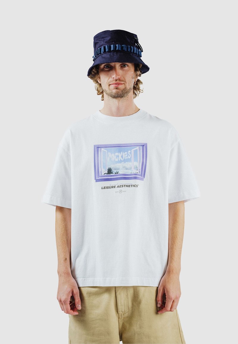 Homme portant un T-shirt blanc avec un motif de fenêtre violet et le texte « LEISURE AESTHETICS », un short beige et un chapeau bob bleu marine.