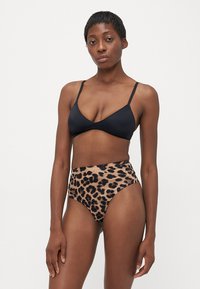 Woman with short hair wearing a black bra and high-waisted leopard print underwear, standing against a plain white background.