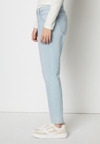 Svetle modré džíny v úzkém střihu, vyrobené z denimové tkaniny s hladkou texturou. Nosené s bílými a béžovými teniskami, zobrazené z boku.