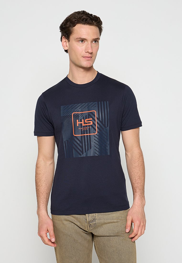 Homme portant un t-shirt graphique bleu marine avec un design en carré orange et noir, pantalon beige, se tenant devant un fond clair uni.