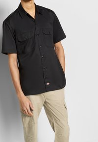 Svart kortärmad skjorta med två framfickor, knäppning och en liten logotypetikett nära fållen, tillverkad av ett mjukt, lätt tyg.