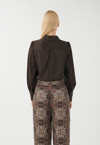 Rjava bluzica s napihnjenimi rokavi, zapenjanjem na zapestjih in naborkom na hrbtu, kombinirana s patterned rjavimi hlačami z okrasnimi motivi.