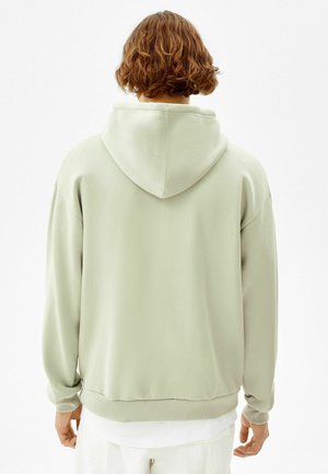 Personne aux cheveux courts et bouclés portant un sweat à capuche vert clair, vue de dos sur un fond blanc uni.