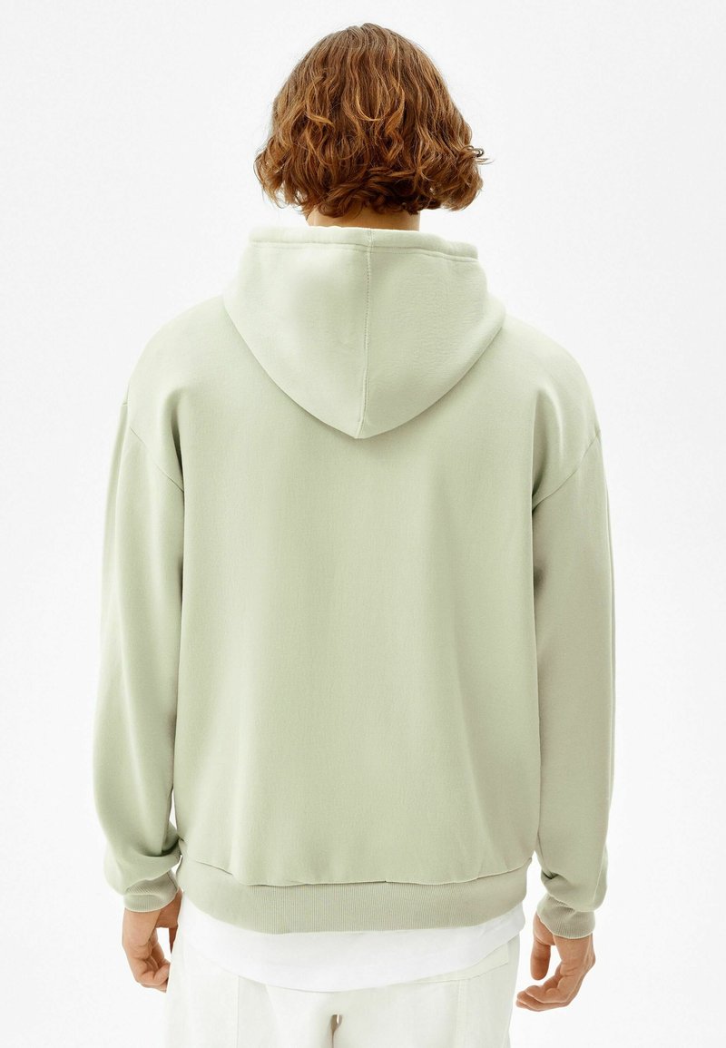 Personne aux cheveux courts et bouclés portant un sweat à capuche vert clair, vue de dos sur un fond blanc uni.