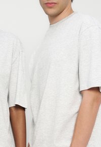 Δύο άτομα φορούν απλά ανοιχτόγρι ζ T-shirt με κοντά μανίκια, εμφανίζονται από τους ώμους μέχρι τη μέση σε λευκό φόντο.