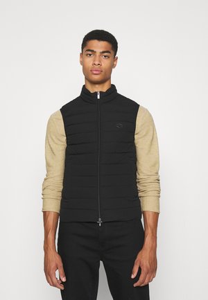 Black padded vest with high collar, front zip closure, and no sleeves, layered over a beige long-sleeve shirt. Simple design.