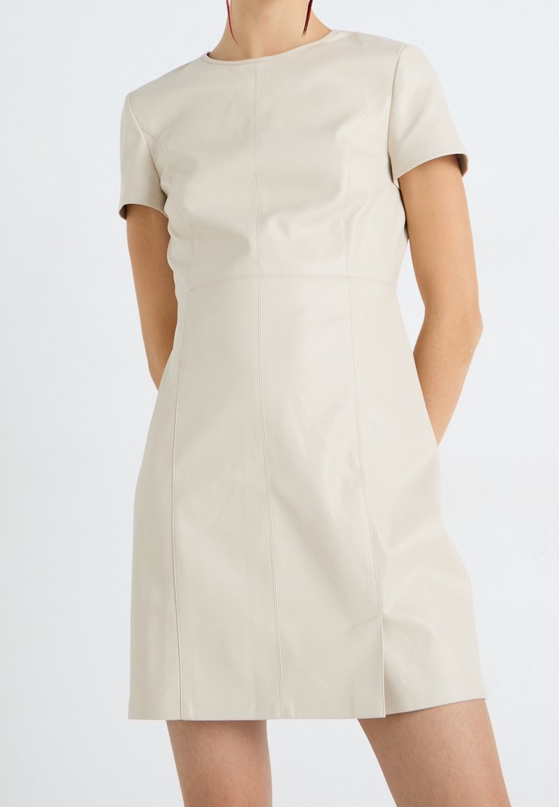 Femme portant une mini robe en cuir beige clair à manches courtes avec des coutures verticales, debout sur un fond blanc uni.