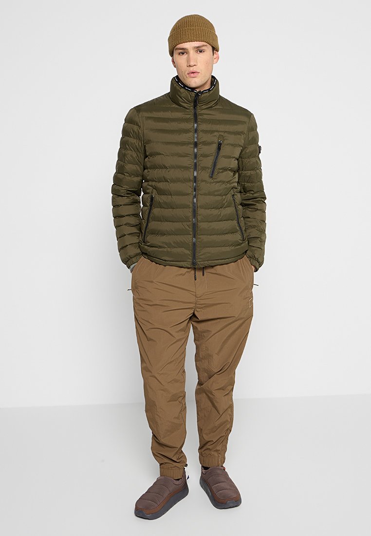 Olijfgroene pufferjas met een hoge kraag, rits aan de voorkant en zijzakken, gecombineerd met beige broek met trekkoord en bruine instappers.