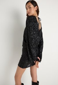 Juoda blizgi suknelė su ilgomis rankovėmis, atvira nugara, uždaryta plonu raiščiu. Turi prigludusią siluetą ir krenta virš kelių.