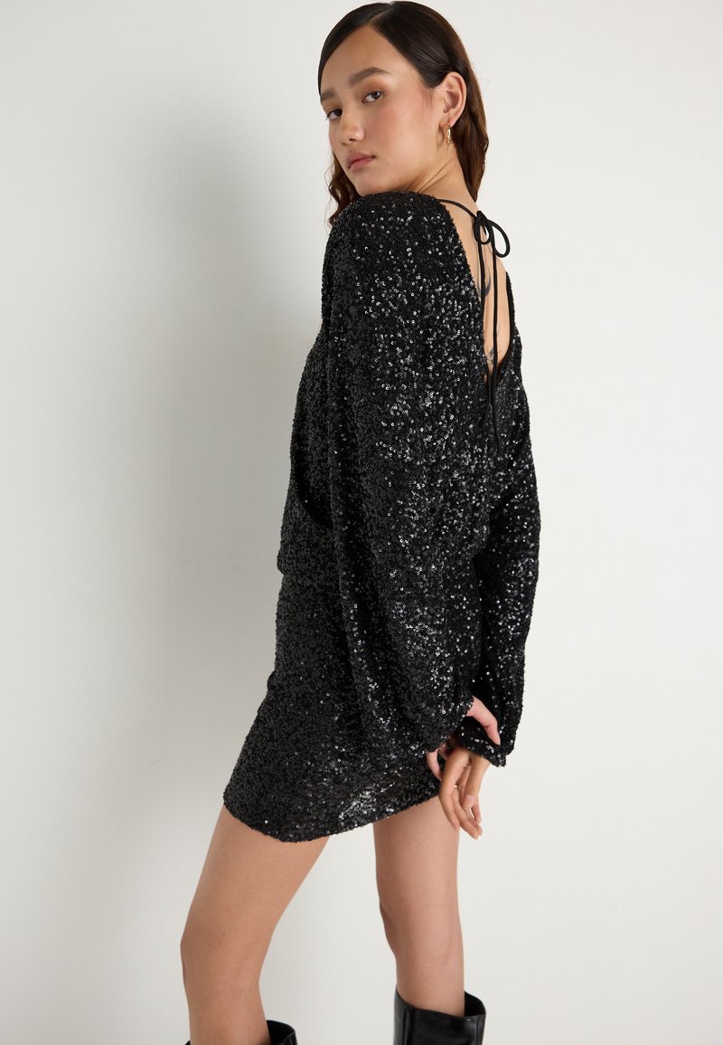 Juoda blizgi suknelė su ilgomis rankovėmis, atvira nugara, uždaryta plonu raiščiu. Turi prigludusią siluetą ir krenta virš kelių.