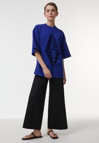 Blusa azul de gran tamaño con un diseño recortado texturizado y en capas, y mangas cortas, combinada con pantalones negros de pierna ancha y sandalias planas.
