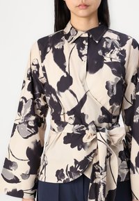 Blusa a fantasia floreale con una combinazione di tonalità crema e scure, chiusura a bottoni, vita legata, maniche ampie e colletto strutturato.