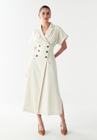 Abito beige fino al ginocchio con maniche corte, design avvolgente, bottoni doppi, tasche laterali e uno spacco alto all'orlo.