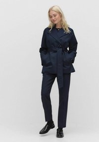 Kobieta w granatowej kurtce z paskiem, w spodniach w prążki i czarnych loafersach, stoi na tle jasnoszarym.