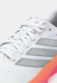 Λευκό αθλητικό παπούτσι με γκρι ρίγες, υφασμάτινο επάνω μέρος με υφή, ροζ εξωτερική σόλα και κορδόνια σε αντίθεση, που παρουσιάζει ένα σπορ σχέδιο.