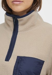 Beige fleece'i päevasoe, kõrge kaelusega ja mereväe sinise tõmblukuga. Omab mereväe sinist tõmbluku taskut rinnal ja tekstuurset kangast. Igapäevane disain.