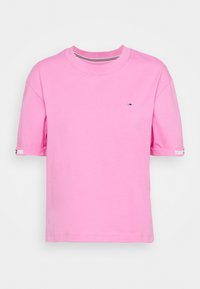 Camiseta rosa de manga corta con pequeño logo de Tommy Jeans en el pecho y la marca Tommy Jeans en blanco en los puños de las mangas.