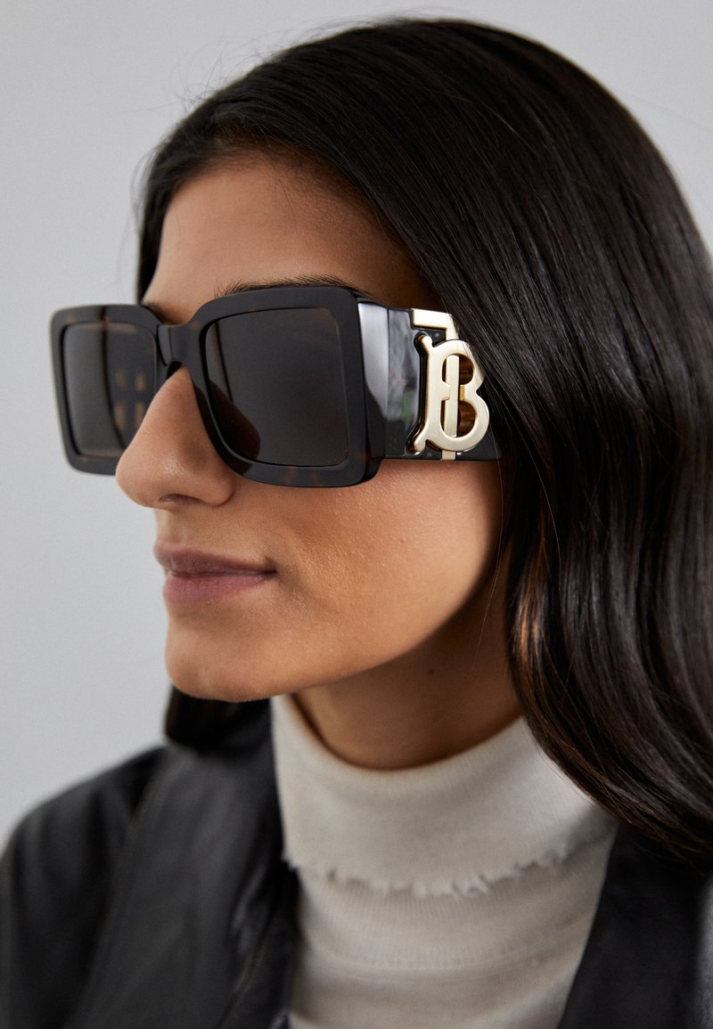 Óculos de sol oversized pretos com um acabamento brilhante, apresentando um accentuador dourado com a letra "B" na lateral e lentes escuras. Detalhe texturizado na armação.