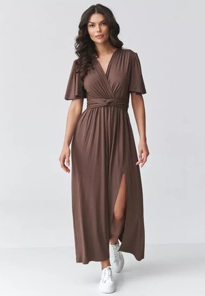 Kobieta z długimi ciemnymi włosami, ubrana w brązową sukienkę kopertową z krótkimi rękawami i przednim rozcięciem, w białych trampkach, stoi na szarym tle.