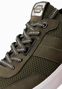 Zapatilla deportiva verde con parte superior de malla y secciones texturizadas. Presenta cordones grisáceos, ojales reforzados y un logo en la lengüeta.
