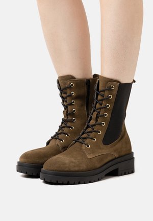 Botas de tornozelo em camurça castanha com painéis laterais elásticos pretos, ilhós dourados e atacadores pretos. Sola de borracha espessa com um padrão de piso robusto.