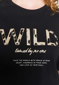 Camiseta de algodón negra con texto en dorado y estampado de leopardo. El texto frontal dice "WILD domesticado por nadie" y hay una cita motivacional abajo.